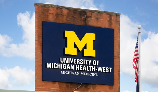 A sign on a brick wall reading "University of Michigan Health-West, Michigan Medicine" beneath a yellow block "M" logo, with an American flag flying beside it against a blue sky.