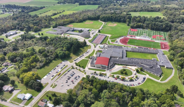 Aerial view of a school campus with multiple buildings, parking lots, athletic fields, tennis courts and a running track surrounded by trees.