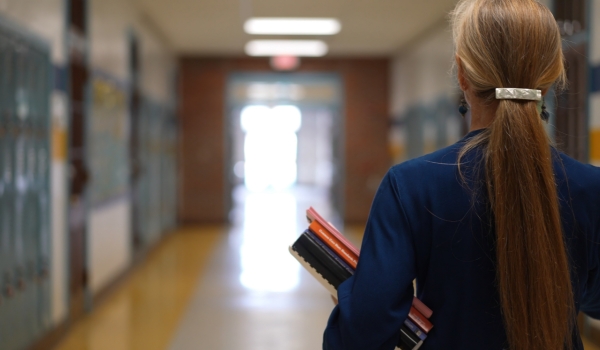Rear view of teacher walking down a hallway in an empty school holding books showing emptiness