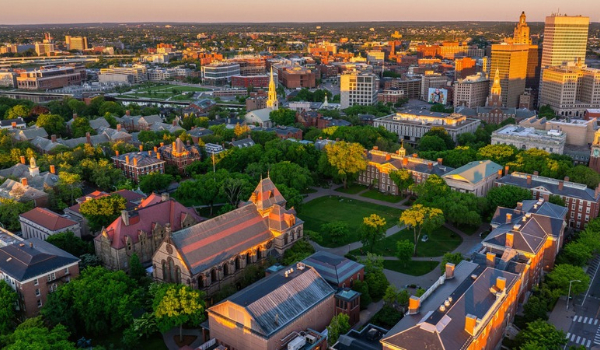 An aerial view at sunset showing the green campus of Brown University in the foreground, with downtown Providence, Rhode Island, skyscrapers in the background.