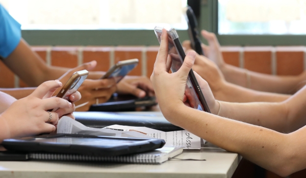 A table of high school students all holding multiple digital devices such as phones and tablets during a class lesson