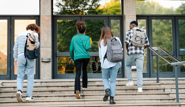 Four students carrying backpacks walk up outdoor stone steps toward the glass doors of a campus building.