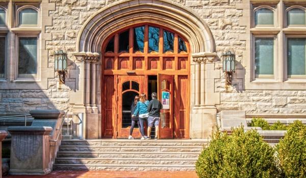 Parents and female student entering large arched doors of University building flanked by metal gothic lamps