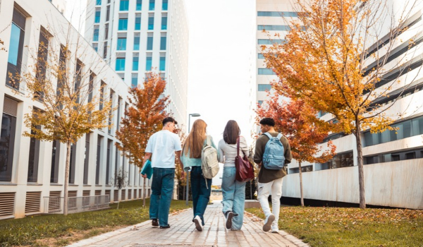 Four students with backpacks walk along a campus pathway between buildings and trees.