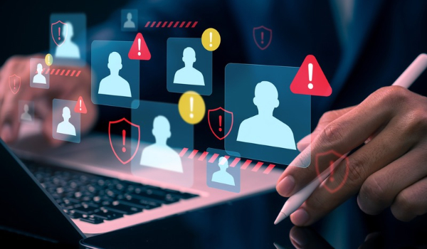 Person using a laptop and stylus with overlaid user profiles, warning symbols and shield icons representing digital threats.