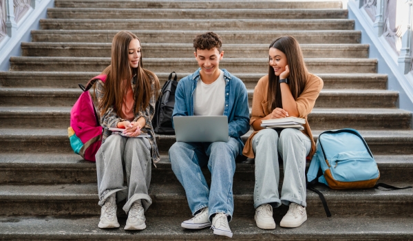Group of high school students sitting on campus stairs with textbooks and laptop, classmates talking together
