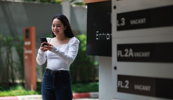 Young Asian woman smiling while looking at her smartphone. Standing outside next to an entrance sign and a directory showing vacant spaces