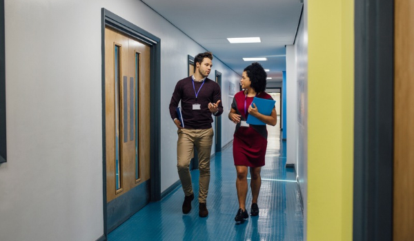 Two colleagues carrying folders talk as they walk down a school hallway.