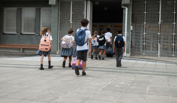 Students wearing uniforms and carrying backpacks walk toward a school entrance.