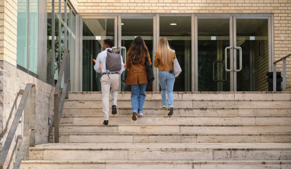 Three university students with backpacks walking up stone steps toward the glass entrance doors of a modern campus building.