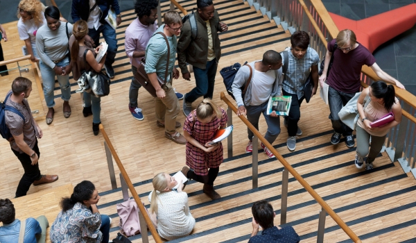 A high-angle view of diverse university students walking up and down a large wooden staircase inside a campus building.