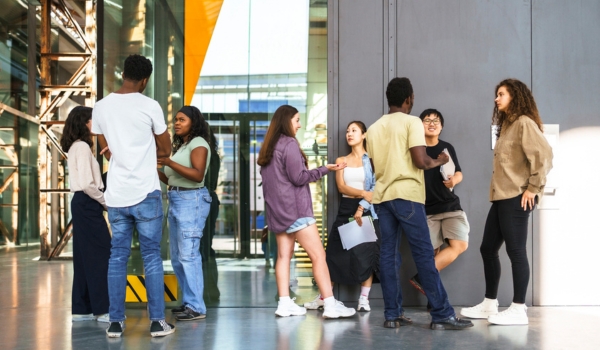 A diverse group of university students stands in small clusters chatting inside a modern campus building.